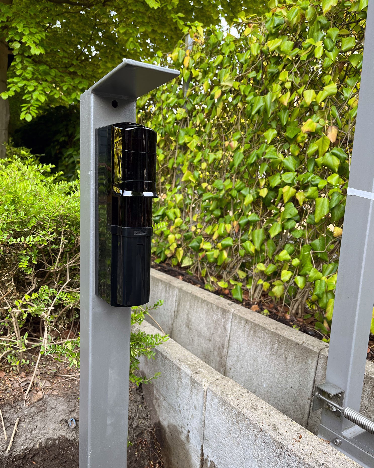 Schwarzer OPTEX-Außenbewegungsmelder auf einem grauen Pfosten am Zaun, davor eine begrünte Hecke.