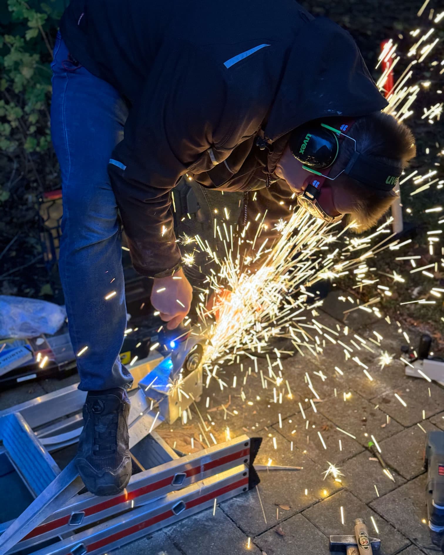 Techniker mit Gehörschutz trennt Metallprofile mit der Trennscheibe, Funkenflug in der Dämmerung.